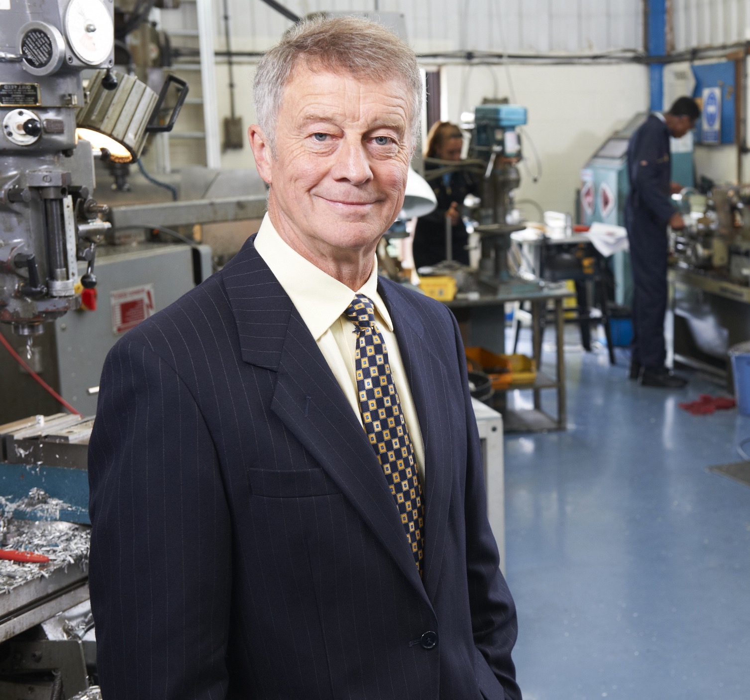 Business owner in a suit standing in his engineering workshop, machinists working at milling machines behind him