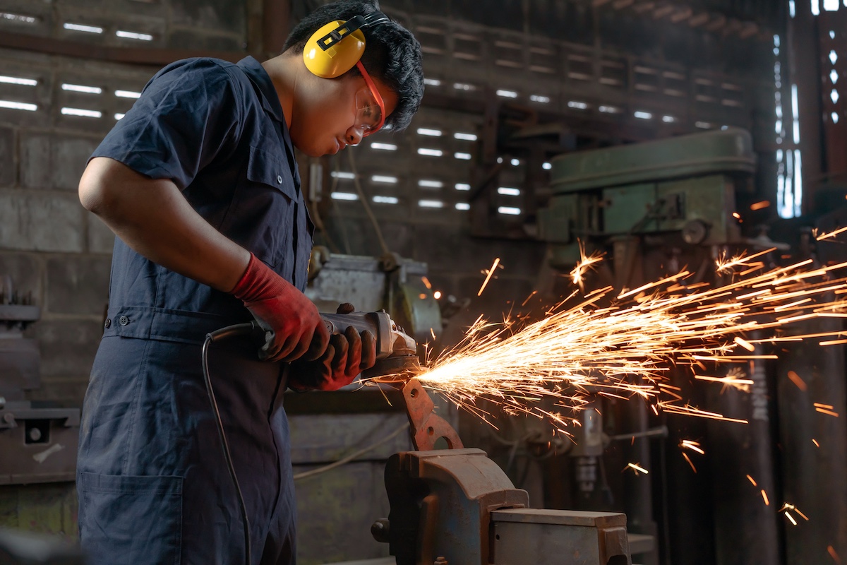 Engineer in overalls, ear defenders and safety goggles cutting a metal bracket with an angle grinder, sparks flying, in a workshop